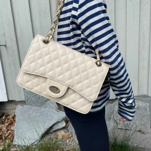Beige handväska  - Säljer en beige handväska som får plats med det mesta med snygg kedjerem och vridspänne i guld. Väskan har klassisk design och är perfekt för att lyfta vilken outfit som helst. Rymmer det viktigaste och är lätt att bära över axeln. Funkar till både fest och skola💘