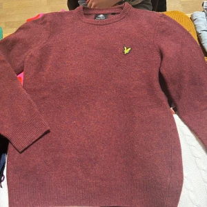 Vinröd tröja från Lyle & Scott - Säljer en snygg vinröd tröja från Lyle & Scott med deras ikoniska logga på bröstet. Tröjan har långa ärmar och en klassisk rund halsringning. Perfekt för kyligare dagar!
