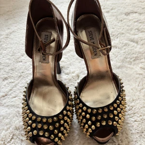 Svarta klackskor med nitar från Steve Madden - Snygga svarta klackskor från Steve Madden med guldnitar och öppen tå. Skorna har en hög klack och en elegant design som passar perfekt för en utekväll. De har en rem runt ankeln för extra stöd.
