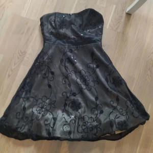 Svart axelbandslös klänning från Miss Etam - Snygg svart axelbandslös klänning från Miss Etam med blommigt broderi och paljettdetaljer. Perfekt för en festlig kväll! Klänningen har en elegant passform och en vacker glansig yta.