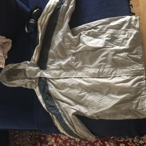 Beige vindjacka från Adidas - Säljer en beige vindjacka från Adidas med huva och dragkedja. Jackan har en klassisk design med Adidas-loggan på sidan. Perfekt för blåsiga dagar och har en bekväm passform. Har gul fläck på bröstet se bild