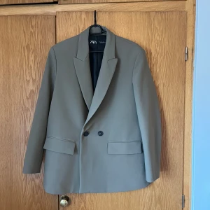 Beige kavaj från Zara - Snygg beige kavaj från Zara med klassisk design och två knappar framtill. Perfekt för en stilren look. Kavajen har långa ärmar och två fickor med lock. Passar bra till både vardag och mer formella tillfällen.