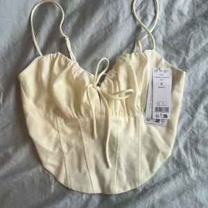 Krämfärgad korsettopp från Bershka - Säljer en stilren krämfärgad (egret) korsettopp från Gina Tricot med smala axelband och knytning fram. Toppen har en elegant design med snörning i ryggen för en justerbar passform. Perfekt för att lyfta din outfit till nästa nivå!