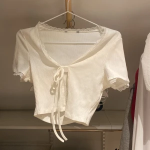 Vit ribbad knytblus - Säljer en söt vit ribbad blus med knytning framtill och spetsdetaljer vid ärmslut och hals. Blusen är kortärmad och croppad, perfekt för sommaren Passar bra till både jeans och kjol!