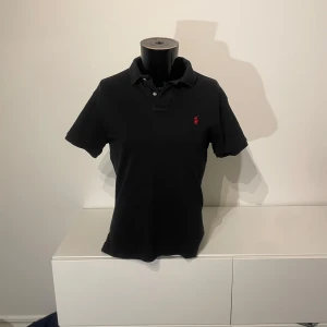 Svart pikétröja med röd logga - Snygg svart pikétröja med en liten röd logga på bröstet. Tröjan har korta ärmar och en klassisk krage med knappar. Perfekt för en stilren look.