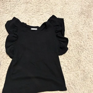 Svart ribbad topp med volangärmar - Snygg svart ribbad topp från Zara med volangdetaljer på axlarna. Perfekt för att ge en extra touch till din outfit. Toppen har en rund halsringning och är ärmlös. Pris går att diskuteras🥰🥰