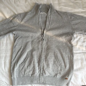 Grå kofta från Mango - Säljer en stilren grå kofta från MNG med dragkedja framtill. Koftan har långa ärmar och ribbade muddar vid ärmslut och nederkant. Perfekt för en avslappnad look. Koftan är i ett bra skick och har ett nypris på ca 250 kr