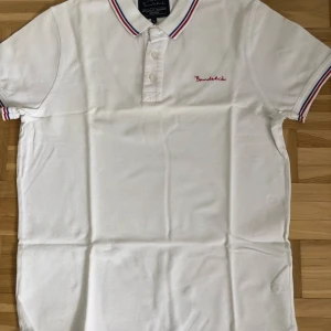 Vit pikétröja från Bondelid - Säljer en stilren vit pikétröja från Bondelid med broderad logga på bröstet. Tröjan har korta ärmar och en klassisk krage med röd och blå randig detalj. Perfekt för en avslappnad och snygg look.