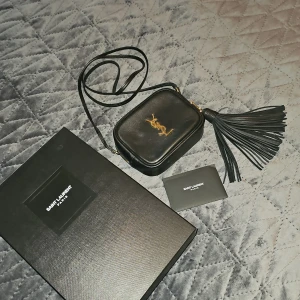 Väska från Saint Laurent / YSL - Camera Lou leather crossbody bag från YSL med guldfärgad logotyp på framsidan. Väskan har en dragkedja och en stor tofsdetalj på sidan. Väskan är lite och behändig med fack på yttersidan samt innersidan. Mått: B14xH10xD6 väskan har box samt kvitto från Inköpsdatum 2018.