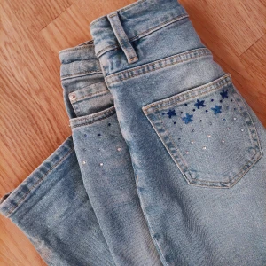 Blå jeans med broderade detaljer - Snygga blå jeans i nyskick med hög midja och raka ben. De har unika broderade blommor och diamanter på bak och framfickor i blått som ger en extra touch. Perfekta för dig som vill ha något annorlunda i garderoben! Materialet är inte super stretchigt men de har en loose fit som gör att de sitter skönt. Hör av dig vid fler frågor eller bilder!💕