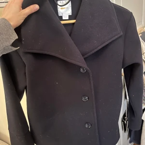 Svart kappa från Gina Tricot - Säljer en stilren mörkblå jacka  från Gina Tricot. Den har en elegant krage och knäpps med knappar framtill. Perfekt för kyligare dagar och ger en klassisk look. Passar bra till både vardag och mer uppklädda tillfällen.anvönd fåtal ggr