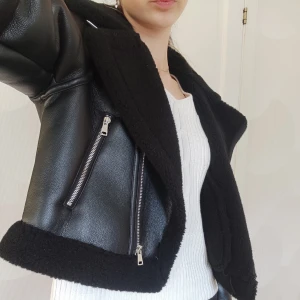 Svart skinnjacka med teddyfoder - Snygg svart skinnjacka med mjukt foder och dragkedjor. Jackan har en cool bikerstil med bred krage och är perfekt för kyligare dagar. Den har långa ärmar och en croppad passform som ger en trendig look.