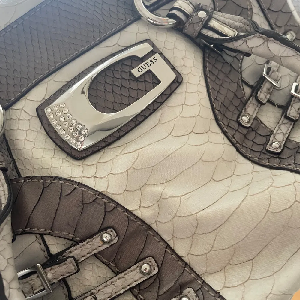 Snygg handväska från Guess i beige och brunt med krokodilmönster. Väskan har dekorativa spännen och en stor metall-logga framtill. . Laukut & Käsilaukut.