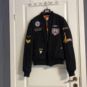 Svart bomberjacka med patchar - Säljer en svart bomberjacka från King of Fashion med coola patchar och broderier. Jackan har dragkedja framtill och ribbade muddar vid ärmslut och nederkant. Insidan är fodrad i orange för en snygg kontrast. Perfekt för dig som vill ha en unik stil! Barn storlek L, som cirka 158.