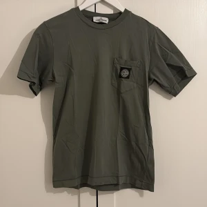 Grön t-shirt från Stone Island Junior - Snygg grön t-shirt från Stone Island Junior med en bröstficka och det ikoniska märket på framsidan. Perfekt för en stilren och avslappnad look. Tröjan har två små fläckar som syns på bild två. Tröjan är inköpt från NK kids i Stockholm.