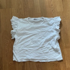 Vit tröja från Zara - Säljer en söt vit tröja från Zara med volangdetaljer på ärmarna. Perfekt för en somrig look. Tröjan har en lös passform och är tillverkad i ett mjukt material.