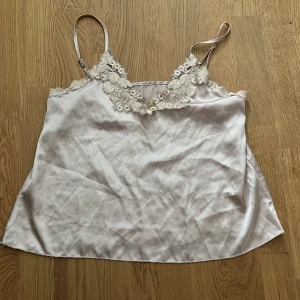 Vit satinlinne med spetsdetaljer - Säljer ett elegant vitt satinlinne med vackra spetsdetaljer vid halsringningen. Linnet har tunna axelband och en lös passform, perfekt för en stilren look. Vet ej vartifrån.