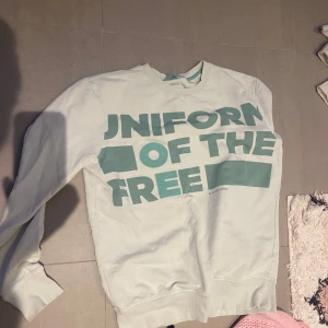 Turkos sweatshirt från G-Star RAW - Säljer en vit sweatshirt från G-Star RAW med texten 'UNIFORM OF THE FREE' i grönt på framsidan. Tröjan har långa ärmar och en klassisk rund halsringning. Perfekt för en avslappnad stil.