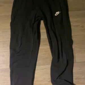 Svarta Nike Byxor - Snygga svarta träningsbyxor från Nike med en vit logga på låret. Perfekta för gymmet eller löprundan. Byxorna har en bekväm passform och är gjorda i ett mjukt material.