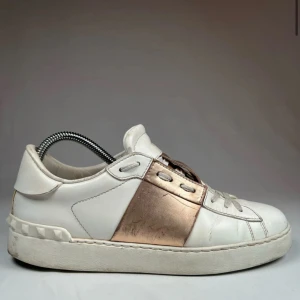 Valentino Garavani sneakers i vitt och guld - Snygga Valentino Garavani sneakers i vitt skinn med en glansig guldpanel över snörningen. Skorna har en klassisk design med rund tå och en bekväm sula. Perfekta för att ge din outfit en lyxig touch.