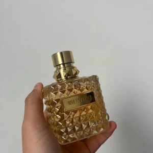 Parfym från Valentino - Elegant parfymflaska från Valentino med ett lyxigt, facetterat mönster i glas och en gyllene kork. Flaskan har en rektangulär form och utstrålar exklusivitet och stil. Perfekt för den som vill ha en doft med både klass och modernitet.