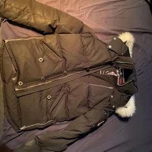 Svart dunjacka med pälsdetaljer - Snygg svart dunjacka med pälsdetaljer på huvan. Jackan har dragkedja och knappar framtill samt flera praktiska fickor. Perfekt för kalla dagar.