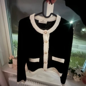 Svart och vit kofta med guldknappar - Jättegullig svart kofta med vita detaljer och guldknappar. Den har långa ärmar och två dekorativa fickor framtill. Perfekt för en stilren look. Passar till allt🥰tröjan är i storlek 146/152 men är lite större i storleken, passar mig som har storlek 158/164❤️Säljer för bra pris!!