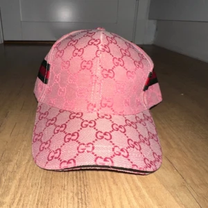 Rosa keps från Gucci - Snygg rosa keps från Gucci med det ikoniska GG-mönstret. Kepsen har gröna och röda ränder på sidorna och justerbar rem baktill. Perfekt för att ge din outfit en lyxig touch.