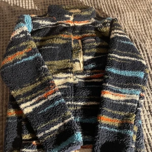 Randig fleecejacka från Columbia - Säljer en mysig fleecejacka från Columbia med randigt mönster i blått, grönt, orange och vitt. Jackan har en dragkedja framtill och är långärmad. Perfekt för kyliga dagar!