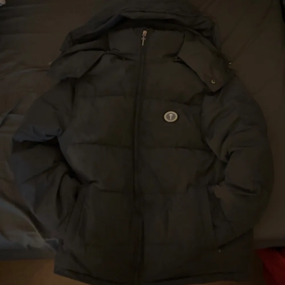 Säljer en svart pufferjacka från Trapstar med dragkedja och huva. Jackan har en stor logga på ryggen och är perfekt för kyligare väder. Den har långa ärmar och en stilren design. Pris kan diskuteras . Takit.