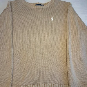 Beige stickad tröja från Ralph Lauren - Säljer en stilren beige stickad tröja från Ralph Lauren med det klassiska broderade logotypen på bröstet. Tröjan har långa ärmar och en rund halsringning. Perfekt för en avslappnad och elegant look.