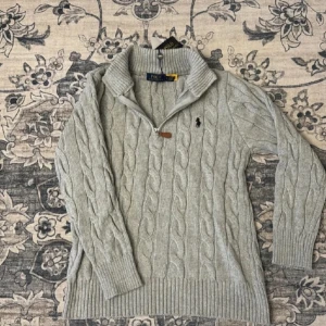 Grå stickad tröja från Ralph Lauren - Säljer en stilren grå stickad tröja från Ralph Lauren med kabelstickat mönster. Tröjan har en dragkedja vid halsen och långa ärmar. Perfekt för kyligare dagar!