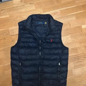 Svart dunväst från Polo Ralph Lauren - Snygg svart dunväst från Polo Ralph Lauren med dragkedja framtill och två fickor med dragkedjor. Den har en diskret röd logga på bröstet. Perfekt för lager-på-lager under kyliga dagar.