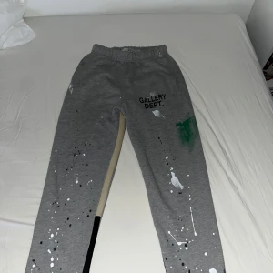 Grå mjukisbyxor från Gallery Dept. - Grå mjukisbyxor från Gallery Dept. med unikt stänkmönster i vitt och grönt. Byxorna har elastisk midja och är tillverkade i bomull för en bekväm passform. Perfekta för en avslappnad stil.