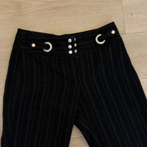 Svarta randiga byxor med detaljer - Snygga svarta byxor med vita ränder och unika metallringar vid midjan. Byxorna har en rak passform och knappar framtill. Perfekta för en stilren look.