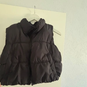 Svart dunväst med dragkedja - Säljer en svart dunväst med dragkedja. Västen har en hög krage och är perfekt för kyligare dagar. Den har en enkel och stilren design med två fickor framtill.