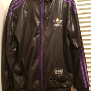 Svart och lila jacka från Adidas - Snygg svart jacka från Adidas med lila detaljer och dragkedja. Jackan har långa ärmar och ett stort Adidas-märke på ryggen. Perfekt för en sportig look.