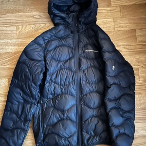 Mörkblå dunjacka från Peak Performance - Säljer en svart dunjacka från Peak Performance. Jackan har en praktisk huva och dragkedja framtill. 