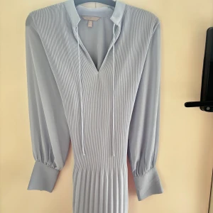 Ljusblå randig klänning från H&M - Säljer en stilren ljusblå klänning från H&M med smala ränder och v-ringning. Klänningen har långa ärmar och knytband vid halsen. Perfekt för en chic look!