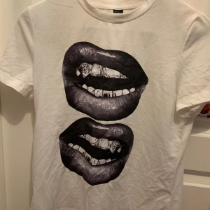 Vit t-shirt med tryck av läppar - Säljer en vit t-shirt med ett coolt tryck av svarta läppar. Perfekt för en avslappnad stil. T-shirten har en normal passform och är kortärmad. Passar bra till jeans eller shorts för en trendig look.