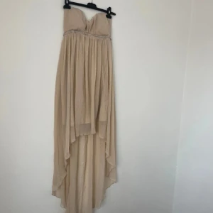 Champagnefärgad axelbandslös balklänning  - Elegant champagnefärgad axelbandslös klänning med vacker fallande design. Klänningen har en glittrig detalj vid bysten som ger en extra touch av glamour. Perfekt för speciella tillfällen. Mer som M än L