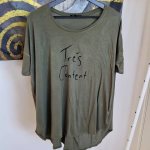 Olivgrön t-shirt från Ellos - Säljer en olivgrön t-shirt från Ellos med texten 'Très Content' tryckt på framsidan. T-shirten har en avslappnad passform och korta ärmar. Perfekt för en casual look!