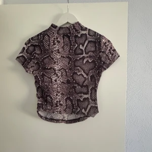 Ormmönstrad kortärmad topp - Säljer en cool kortärmad topp med ormmönster i svart och grått. Perfekt för en trendig look. Toppen har en hög hals och är tillverkad i ett lite genomskinligt material .