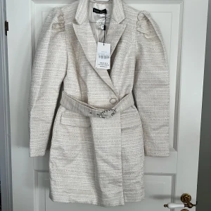 Kavaj klänning från Lavish Alice ny med prislapp  - Elegant klänning från Lavish Alice med ett bälte som har en glittrande spänne. Kappan har en strukturerad yta och långa ärmar med puffiga axlar. Perfekt för en stilren look. Ny med prislapp går även att använda som kappa 