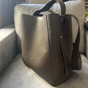 Grå axelväska från Gina Tricot - Snygg grå axelväska från Gina Tricot i syntetmaterial. Väskan har en stilren design med dragkedja och knäppning upptill. Perfekt för att bära med sig det nödvändigaste med stil. Köpt för 550kr