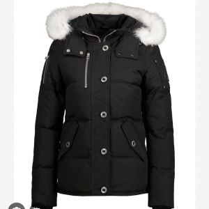 Svart dunjacka med pälsdetaljer - Snygg svart dunjacka med vit päls på huvan. Jackan har dragkedja och knappar framtill samt flera praktiska fickor. Perfekt för kalla vinterdagar. (Små i storlek), (kvitto finns, köpt i Johnells för 10,000kr)