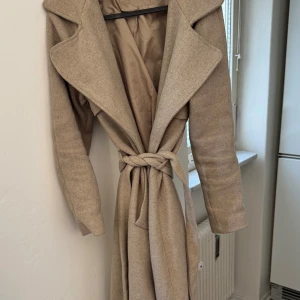 Beige kappa från Bik Bok - Säljer en stilren beige kappa från Bik Bok. Den har en elegant krage och knyts i midjan med ett bälte. Perfekt för höst och vår med sin långa design och långa ärmar. En klassisk och tidlös look för alla tillfällen.