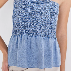 Blå axelbandslös topp - Säljer en söt blå axelbandslös topp med smockdetaljer och volangkant. Perfekt för sommardagar och kan enkelt matchas med olika outfits. Toppen har en luftig och bekväm passform.