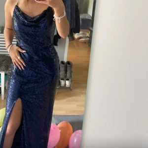 Mörkblå glittrig bal klänning  - Snygg mörkblå klänning med glittrande paljetter och en elegant slits framtill. Klänningen har en draperad halslinje och är ärmlös, perfekt för festliga tillfällen som till exempel bal . Har en lite mer öppen rygg med korsande band 