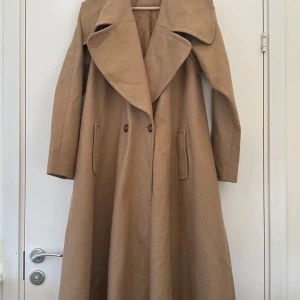 Beige kappa från ASOS - Superfin beige lång kappa från ASOS i storlek 40. Bred klassisk krage och fin dubbelknäppning fram. Elegant klockformad vidd nedtill som gör alla kroppsformer smickrande. Snygg både som den är eller med skärp i midjan.   Nästan nyskick (aldrig använd, men stått i garderoben). Älskar den, men måste kbt:a mig igenom mitt hoardande och ge den en bättre ägare.  😅  Hämtas i Stockholm eller skickas mot porto. 